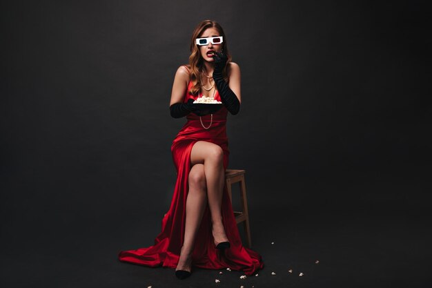 Donna in abito lungo rosso guarda film spaventoso e mangia popcorn Ritratto di signora in abito di raso che posa in occhiali 3d su sfondo nero