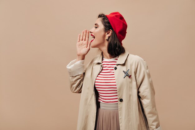 Donna in berretto rosso e trincea grida su sfondo beige. Ragazza alla moda con labbra luminose in maglione a righe e pose cappotto alla moda per la fotocamera.