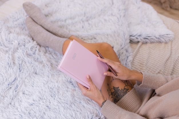 Donna in calza di lana e maglione con taccuino rosa segno 2021, grande tatuaggio sul fianco. La donna si siede sul letto a casa in camera da letto.