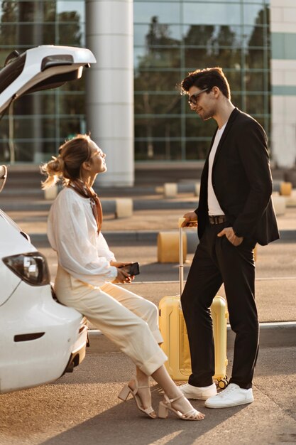 Donna in camicetta bianca pantaloni beige si siede nel bagagliaio dell'auto e parla con il suo fidanzato L'uomo bruna e la signora bionda parlano e posano con la valigia gialla vicino all'aeroporto