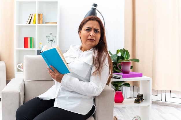 Donna in camicia bianca e pantaloni neri che legge un libro con una faccia seria mentre è seduta sulla sedia in un soggiorno luminoso