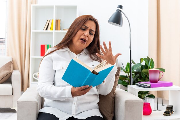 Donna in camicia bianca e pantaloni neri che legge un libro dall'aspetto emotivo e incuriosito seduta sulla sedia in un soggiorno luminoso