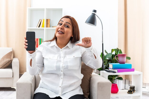 Donna in camicia bianca e pantaloni neri che mostra lo smartphone che punta con il dito indice sorridendo seduto sulla sedia in un soggiorno luminoso