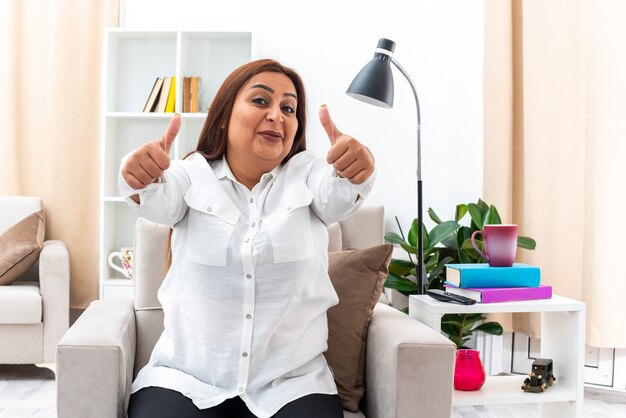 Donna in camicia bianca e pantaloni neri che sembra felice e allegra che mostra i pollici seduta sulla sedia in un soggiorno luminoso