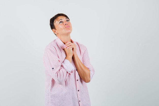 Donna in camicia rosa stringendo le mani nel gesto di preghiera e guardando impotente, vista frontale.