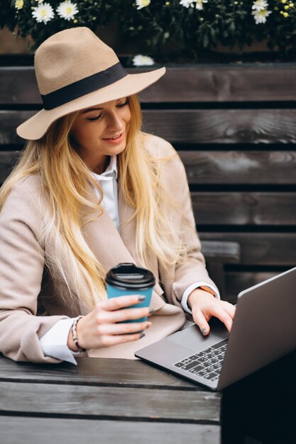 Donna in cappello che beve caffè e che lavora al computer portatile all&#39;esterno