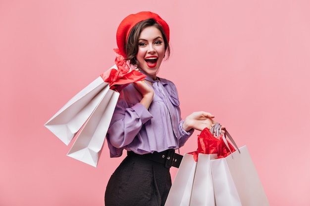 Donna in cappello rosso, pantaloni neri e camicetta leggera ride e posa con i pacchetti dopo lo shopping.