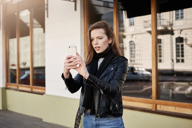 donna in cappotto di pelle alla moda che tiene smartphone mentre si scatta foto di paesaggi o band che suona in strada