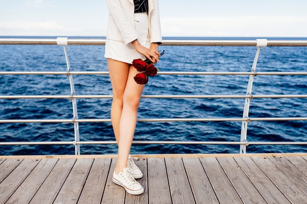 Donna in elegante abito bianco minimalista e occhiali da sole rotondi retrò, con piccole rose rosse