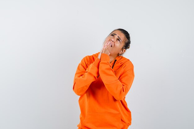 Donna in felpa con cappuccio arancione che tiene le mani in un gesto di preghiera e sembra speranzosa