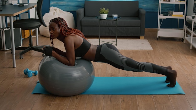 Donna in forma atletica che lavora i muscoli addominali mentre è seduta su una palla svizzera di yoga durante l'allenamento mattutino in soggiorno praticando l'aerobica. Fit adulto che indossa abbigliamento sportivo facendo posizione di aerobica