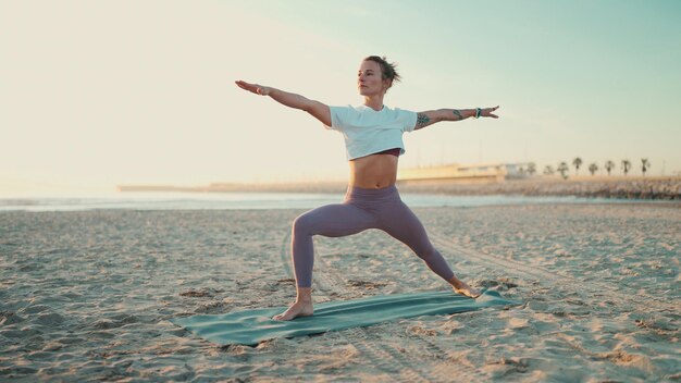 Donna in forma che fa yoga sul materassino in riva al mare Bella ragazza yogi in abbigliamento sportivo in piedi in posa yoga guerriero sulla spiaggia