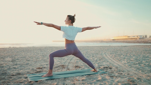 Donna in forma che fa yoga sul tappetino all'aperto Ragazza yogi attraente in abbigliamento sportivo che sembra buono in piedi in posa yoga guerriero sulla spiaggia