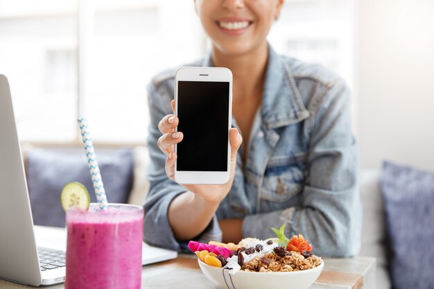 Donna in giacca di jeans alla moda in caffetteria con schermo dello smartphone in bianco