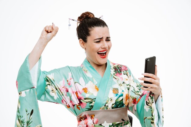 Donna in kimono giapponese tradizionale che tiene il pugno serrato dello smartphone esultando per il suo successo felice ed eccitata su bianco