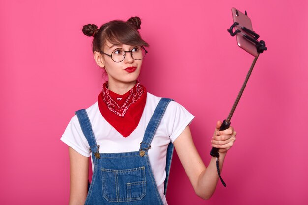 Donna in maglietta bianca casual, tuta, bandana rossa sul collo e occhiali rotondi. Adorabile adolescente mantiene le labbra arrotondate