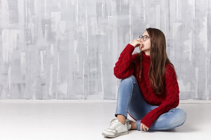 Foto gratuito donna in maglione accogliente lavorato a maglia, occhiali da vista, jeans e scarpe da ginnastica che si siede sul pavimento e distoglie lo sguardo con espressione facciale pensierosa, pensando a problemi adolescenziali
