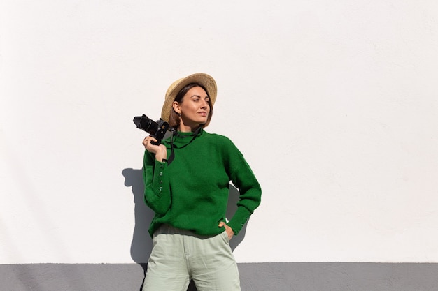 Donna in maglione casual verde e cappello all'aperto sul turista positivo felice muro bianco con telecamera professionale