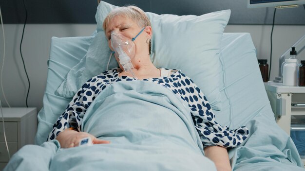 Donna in pensione con tubo dell'ossigeno contro il problema respiratorio nel letto del reparto ospedaliero. Paziente malato che respira pesantemente mentre riposa e in attesa di assistenza medica con sacca per flebo IV.