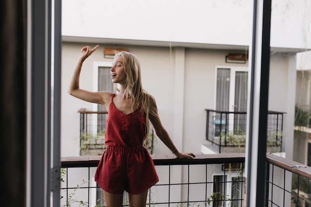 donna in pigiama in piedi sul balcone e guardandosi intorno. Modello femminile emozionante con capelli biondi lunghi che gode del mattino.