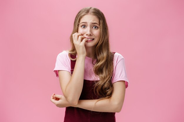 Donna in preda al panico e reazione eccessiva sensazione nervosa che si morde le unghie e fissa impaurita la telecamera tremante di paura su sfondo rosa che posa in graziose salopette con acconciatura ondulata naturale.