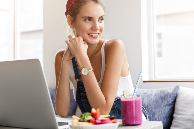 Donna in tuta di jeans alla moda con il computer portatile nella caffetteria