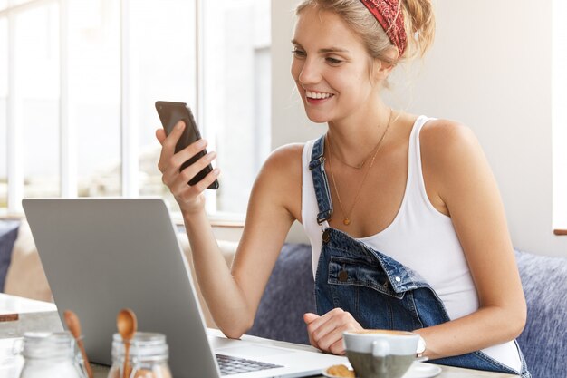 Donna in tuta di jeans alla moda con il computer portatile nella caffetteria