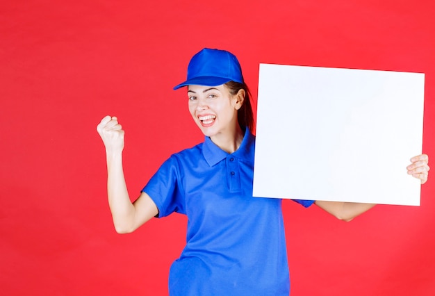 Donna in uniforme blu e berretto che tiene in mano un banco informazioni quadrato bianco e mostra un segno di successo con la mano.