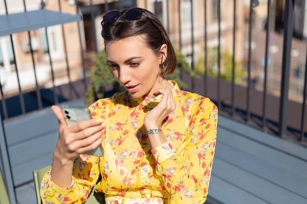 Donna in vestito giallo sulla terrazza in caffè estivo con il telefono cellulare in giornata di sole prendere selfie effettuare una videochiamata