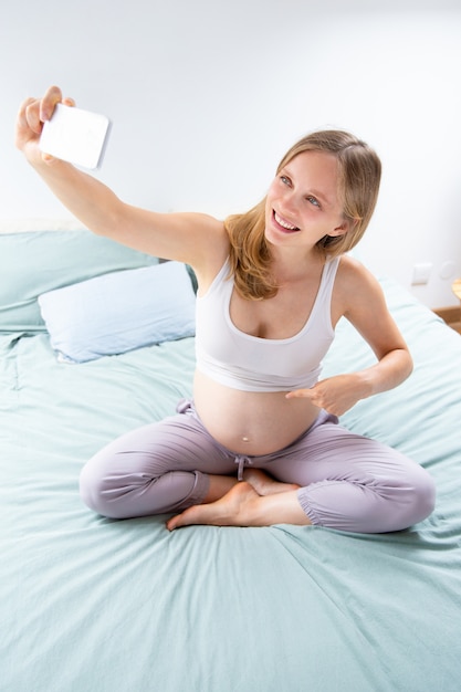 Donna incinta allegra felice che prende selfie in camera da letto