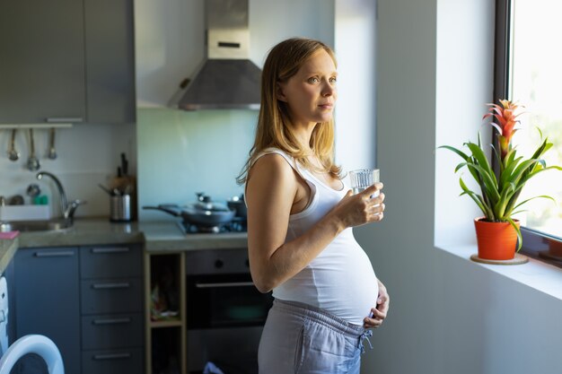 Donna incinta pensierosa positiva che sta nella cucina