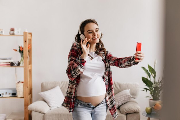 Donna incinta riccia bruna in camicia a quadri prende selfie e ascolta musica in cuffie bianche Madre felice in jeans tiene il telefono rosso e sorride in soggiorno