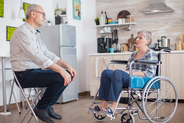 Donna invalida in pensione in sedia a rotelle che ha una conversazione con il vecchio marito anziano in cucina. Vecchio che parla con la moglie. Vivere con una persona disabile con disabilità motorie