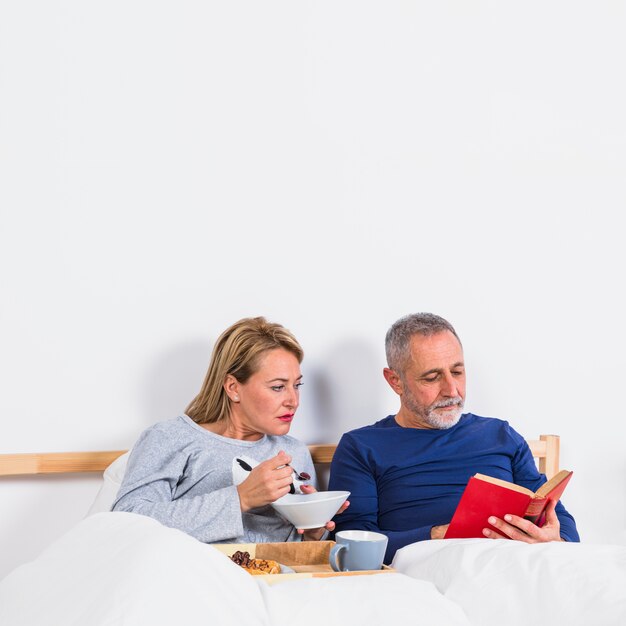 Donna invecchiata con la ciotola vicino all&#39;uomo con il libro in piumino vicino a colazione sul vassoio sul letto