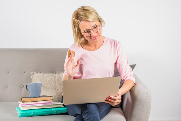 Donna invecchiata in camicia rosa con carta di plastica, laptop e tazza sul mucchio di libri sul divano