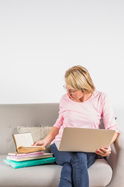 Donna invecchiata in camicia rosa con laptop e mucchio di libri sul divano