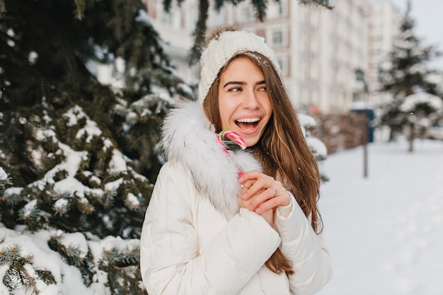 Donna inverno gioiosa divertente con lecca-lecca in città. Divertirsi con la neve, umore pazzo, sorridenti, emozioni positive e luminose. Nuovo anno in arrivo, tempo freddo, tempo felice.