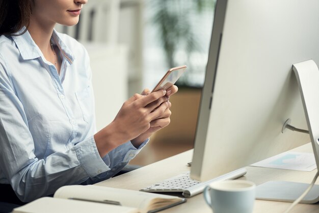 Donna irriconoscibile che si siede nell'ufficio davanti al computer e che per mezzo dello smartphone