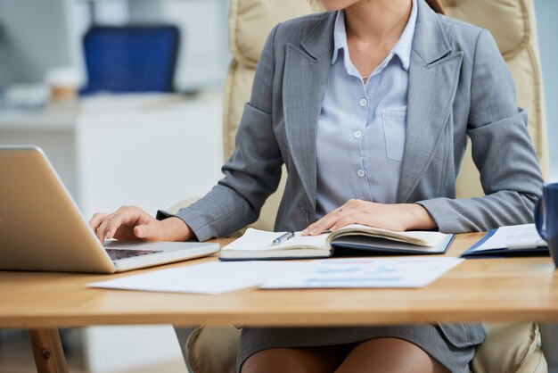 Donna irriconoscibile in vestito che si siede allo scrittorio e che lavora al computer portatile