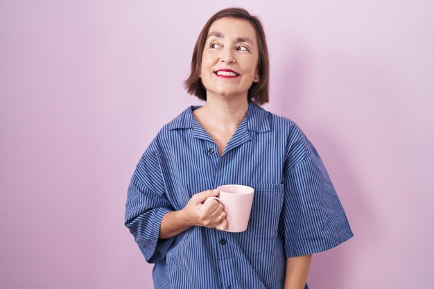 Donna ispanica di mezza età che beve una tazza di caffè sorridente guardando di lato e fissando via il pensiero.