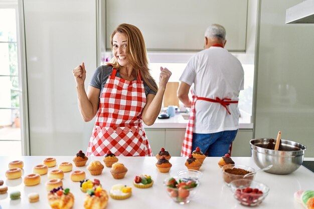 Donna ispanica di mezza età che indossa un grembiule che cucina dolci fatti in casa urlando orgogliosa, celebrando la vittoria e il successo molto eccitati con le braccia alzate