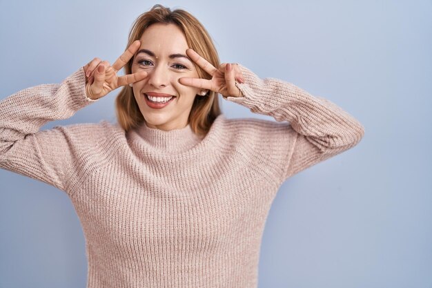 Donna ispanica in piedi su uno sfondo blu che fa un simbolo di pace con le dita sul viso sorridendo allegra mostrando la vittoria