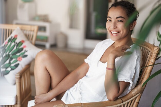 Donna ispanica sorridente felice che si siede sulla poltrona del rattan sul patio dell'hotel