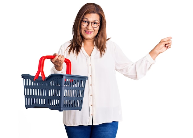 Donna latina di mezza età che tiene il carrello della spesa del supermercato sorridendo felice indicando con la mano e il dito di lato