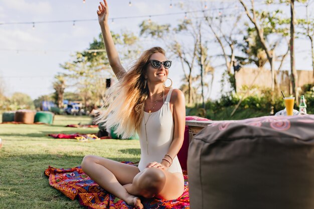 Donna leggermente abbronzata in costume da bagno seduto sul prato e agitando la mano. Outdoor ritratto di bella giovane donna con capelli biondi divertendosi nel parco.