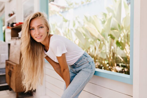 Donna leggermente abbronzata interessata in posa nella mattina di sole. Foto dell'interno della ragazza bionda alla moda in piedi vicino alla finestra.