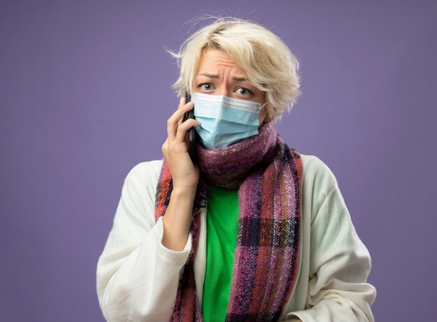 Donna malata malsana con i capelli corti in sciarpa calda e maschera protettiva facciale che sembra stressata e preoccupata mentre parla al telefono cellulare su sfondo viola