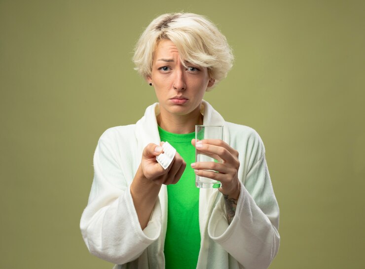 Foto gratuito donna malata sconvolta con capelli corti sensazione di malessere tenendo un bicchiere di acqua e blister con pillole guardando con espressione triste in piedi sopra la parete chiara