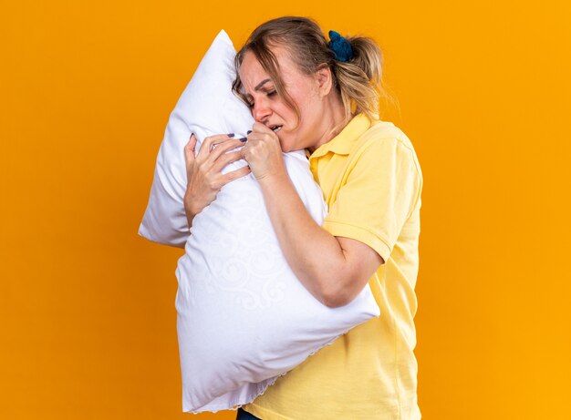 Donna malsana in camicia gialla che si sente male soffre di influenza e cuscino freddo che abbraccia tosse in piedi sul muro arancione