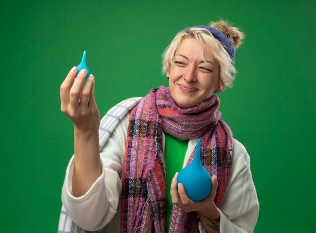 Donna malsana malata con capelli corti in sciarpa calda e cappello avvolto in una coperta che tiene i clisteri guardandolo sorridente in piedi sopra il muro verde